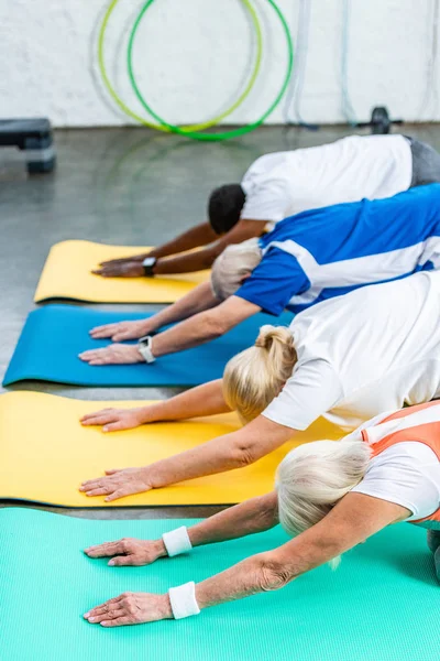 Vista lateral de atletas sénior multiculturales haciendo ejercicio síncrono en colchonetas de fitness en el gimnasio - foto de stock