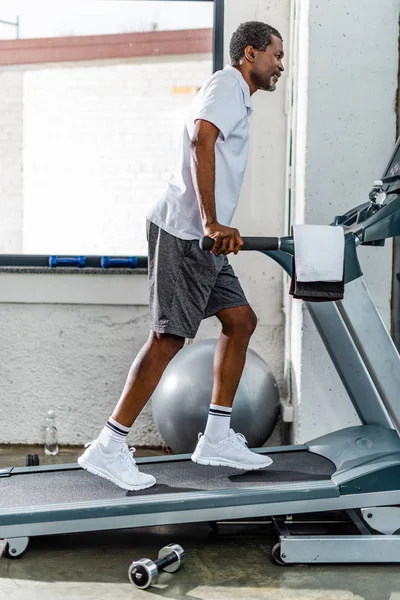 Vista lateral del hombre afroamericano corriendo en la cinta en el gimnasio - foto de stock