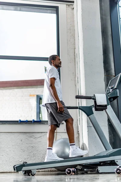 Vista de ángulo bajo del hombre afroamericano de mediana edad haciendo ejercicio en la cinta de correr en el gimnasio - foto de stock