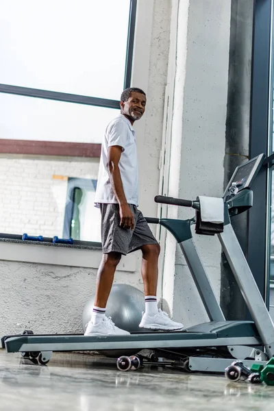 Vista basso angolo di sorridente uomo africano americano che esercita sul tapis roulant in palestra — Foto stock