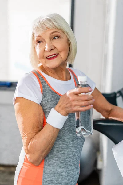 Enfoque selectivo de deportista senior alegre sosteniendo botella de agua y descansando cerca de la cinta de correr en el gimnasio - foto de stock