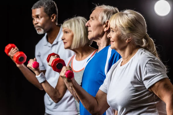 Vista laterale degli sportivi senior mutlietnici sincroni che si esercitano con i manubri su nero con riflettori — Foto stock