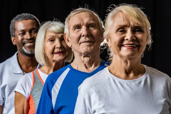 Smiling multicultural senior sportspeople standing isolated on black — Stock Photo