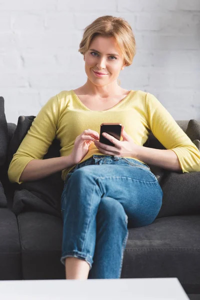 Hermosa mujer usando teléfono inteligente y sonriendo a la cámara - foto de stock