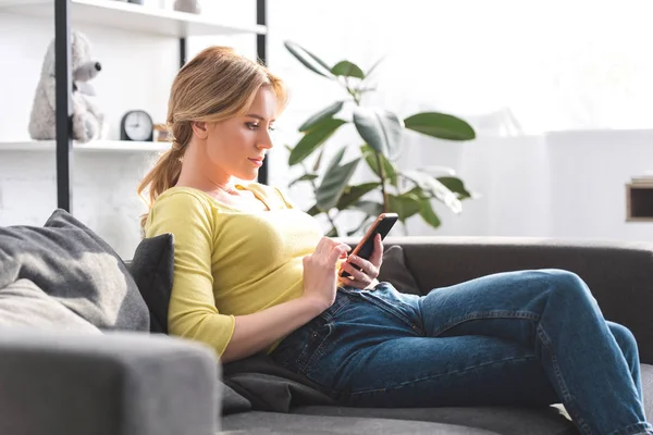 Vista lateral da bela mulher sentada no sofá e usando smartphone em casa — Fotografia de Stock