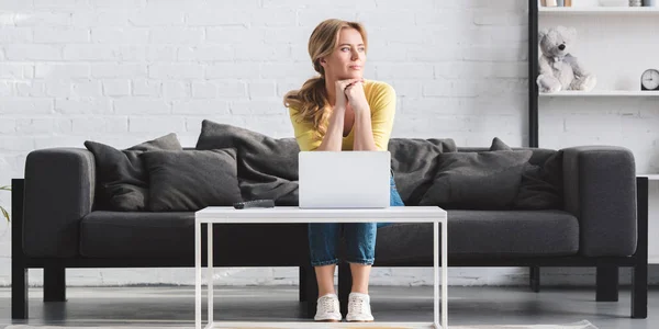 Schöne nachdenkliche Frau, die wegschaut, während sie auf der Couch sitzt und zu Hause Laptop benutzt — Stockfoto