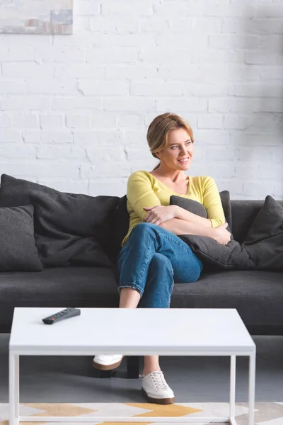 Schöne lächelnde Frau, die auf der Couch sitzt und zu Hause wegschaut — Stockfoto