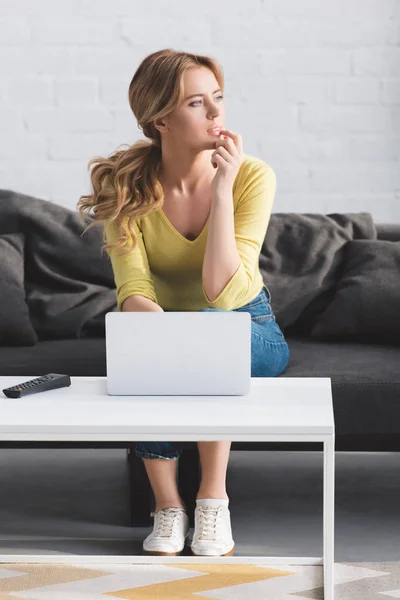 Mujer pensativa mirando hacia otro lado mientras está sentado en el sofá y el uso de la computadora portátil - foto de stock