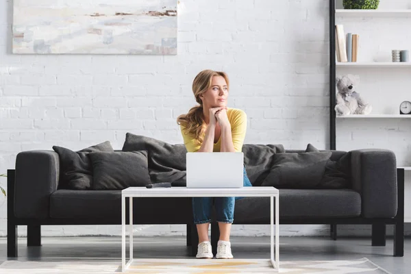 Hermosa mujer mirando hacia otro lado mientras está sentado en el sofá y el uso de la computadora portátil en casa - foto de stock