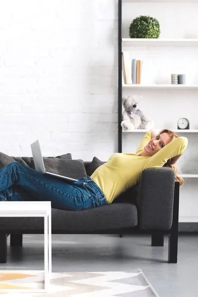 Belle femme heureuse couchée sur le canapé avec ordinateur portable et souriant à la caméra — Photo de stock