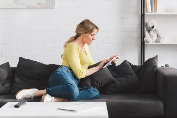 Усміхнена жінка сидить на дивані і використовує цифровий планшет вдома — стокове фото