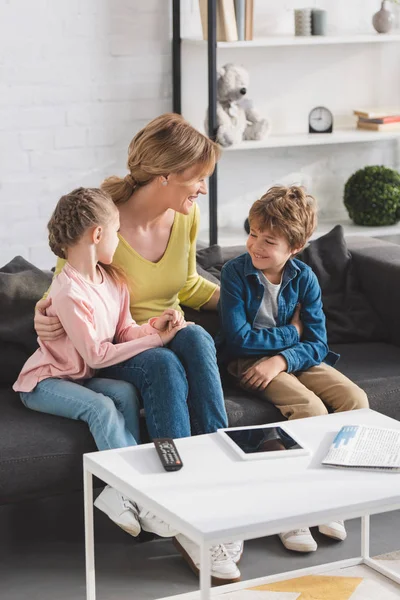 Vista de ángulo alto de la madre feliz mirando a los niños sonrientes adorables sentados juntos en el sofá - foto de stock