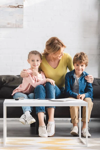 Счастливая мать смотрит на очаровательных улыбающихся детей, сидящих вместе на диване — стоковое фото