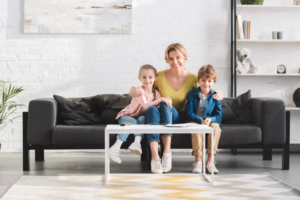 Счастливая мать с восхитительными детьми, сидящими на диване и улыбающимися на камеру — стоковое фото