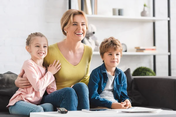 Madre alegre y hermosos niños sonrientes sentados en el sofá y viendo la televisión en casa - foto de stock