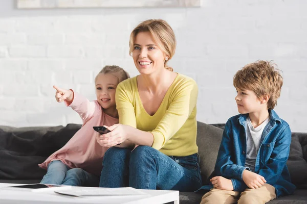 Madre feliz con los niños sonrientes usando el mando a distancia y viendo la televisión juntos - foto de stock