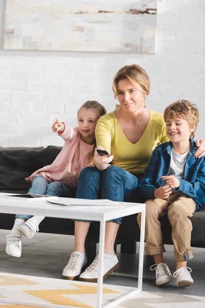 Glückliche Mutter mit entzückend lächelnden Kindern, die Fernbedienung benutzen und gemeinsam fernsehen — Stockfoto