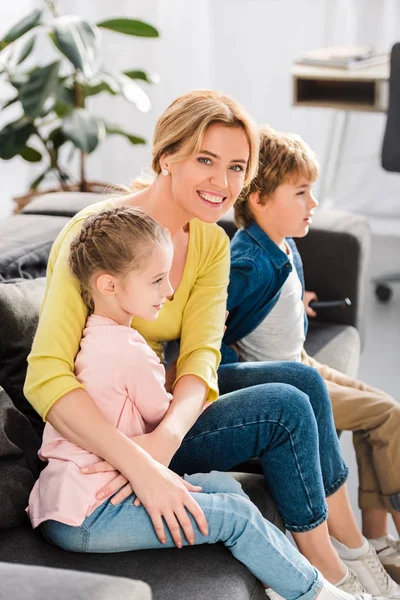 Mère heureuse souriant à la caméra tout en regardant la télévision avec des enfants à la maison — Photo de stock