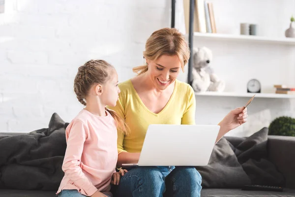 Happy mother holding credit card and using laptop with little daughter at home — Stock Photo
