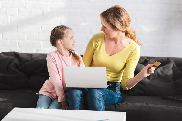 Mãe feliz e filha sorrindo uns aos outros enquanto faz compras on-line com laptop e cartão de crédito — Fotografia de Stock