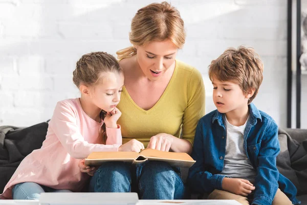 Счастливая мать с милыми маленькими детьми, сидящими на диване и читающими вместе книгу — стоковое фото