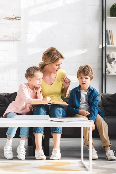 Mãe com filhos adoráveis sentados no sofá e ler livro juntos em casa — Fotografia de Stock