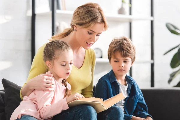 Smiling mother with adorable children reading book together at home — Stock Photo