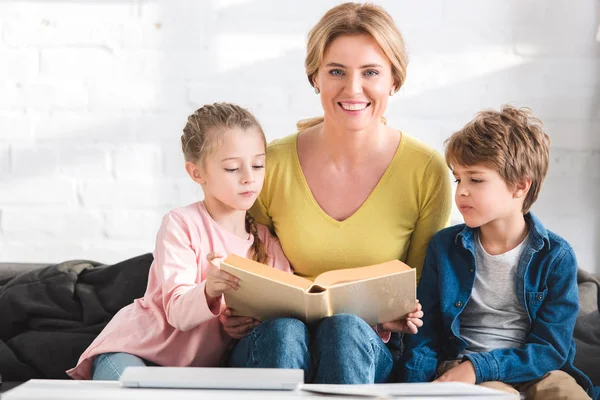 Glückliche Mutter lächelt in die Kamera, während sie mit entzückenden Kindern zu Hause Buch liest — Stockfoto