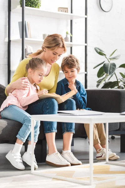 Mère avec des enfants adorables assis sur le canapé et le livre de lecture — Photo de stock