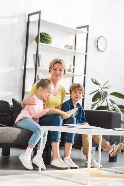 Glückliche Mutter lächelt in die Kamera, während sie mit Kindern zu Hause Buch liest — Stockfoto