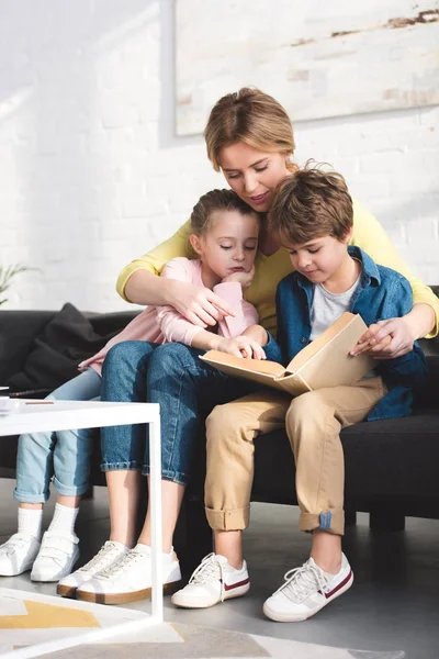 Улыбающаяся мама с милыми маленькими детьми, сидящими на диване и читающими книгу — стоковое фото
