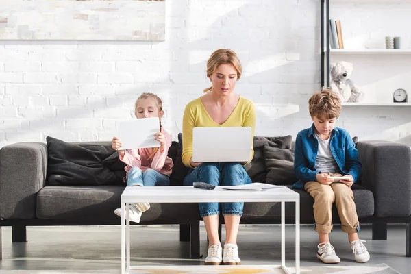 Мать и красивые дети сидят на диване и с помощью цифровых устройств — стоковое фото