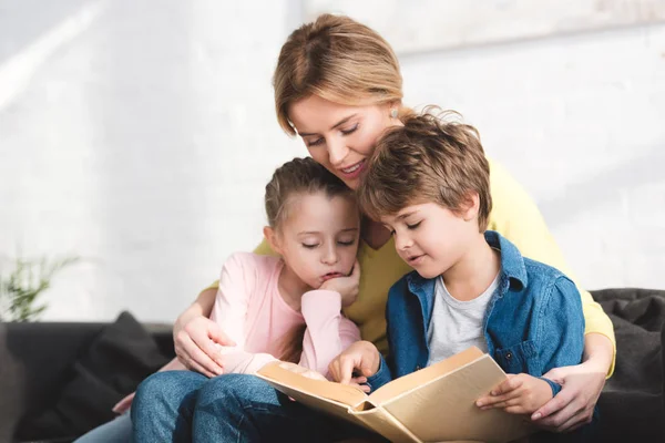 Mère avec des petits enfants mignons assis sur le canapé et le livre de lecture — Photo de stock