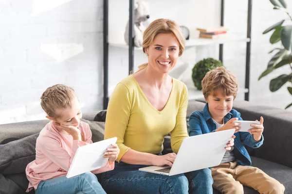 Madre felice usando il computer portatile e sorridendo alla fotocamera mentre i bambini utilizzano dispositivi digitali a casa — Foto stock