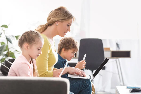 Vista laterale della madre con bambini adorabili che utilizzano dispositivi digitali a casa — Foto stock