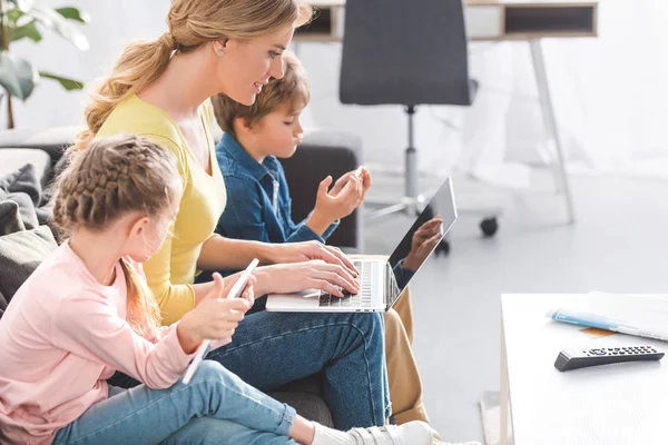 Vista laterale di madre sorridente con bambini adorabili che utilizzano dispositivi digitali a casa — Foto stock