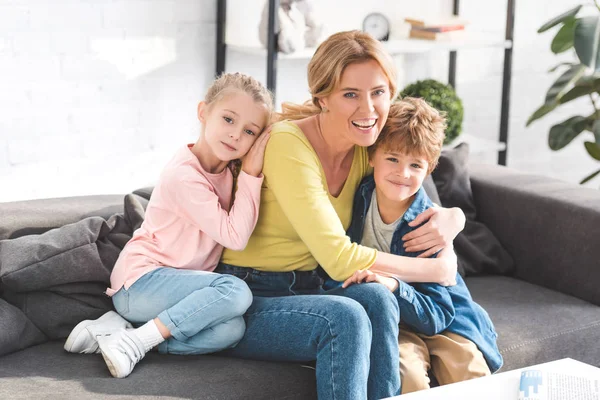 Feliz madre y adorables niños pequeños sentados en el sofá y sonriendo a la cámara - foto de stock