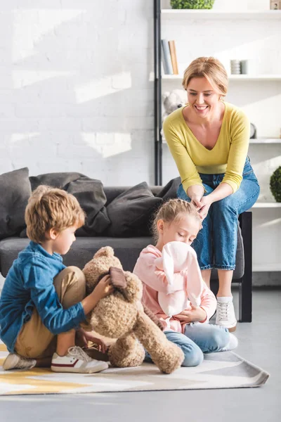 Счастливая мама сидит на диване и смотрит на очаровательных улыбающихся братьев и сестер, играющих с игрушками дома — стоковое фото