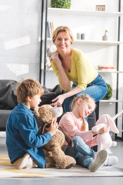 Glückliche Mutter lächelt in die Kamera, während süße kleine Kinder zu Hause mit Spielzeug spielen — Stockfoto