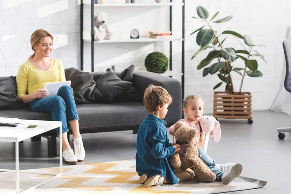 Glückliche Mutter nutzt digitales Tablet und betrachtet entzückende Kinder, die mit Spielzeug spielen — Stockfoto