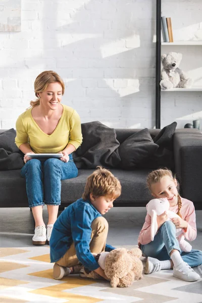 Happy mother holding digital tablet and looking at adorable children playing with toys — Stock Photo