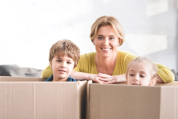 Happy mother smiling at camera and cute little children sitting in cardboard boxes — Stock Photo
