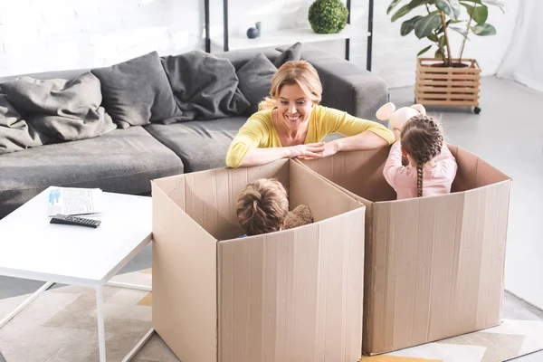 Vista de ángulo alto de la madre feliz mirando a los niños pequeños y lindos jugando en cajas de cartón - foto de stock