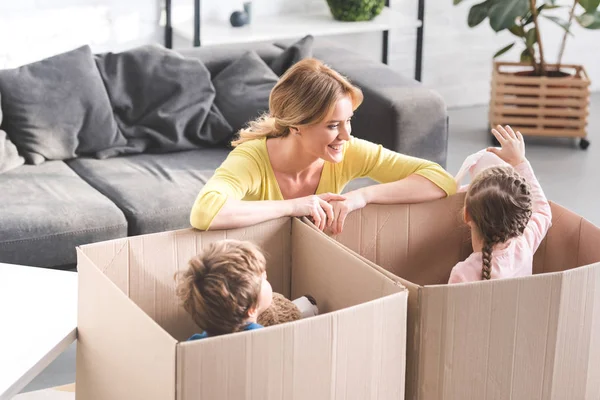 Mère heureuse regardant de mignons petits enfants jouer dans des boîtes en carton — Photo de stock