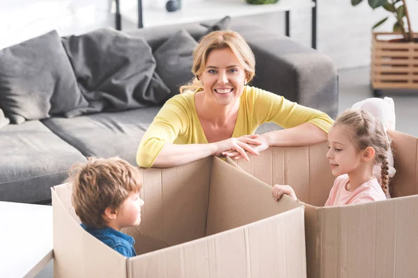 Feliz madre sonriendo a la cámara mientras los niños juegan en cajas de cartón - foto de stock