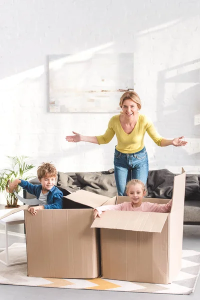 Feliz madre sonriendo a la cámara mientras juega con los niños sentados en cajas de cartón - foto de stock