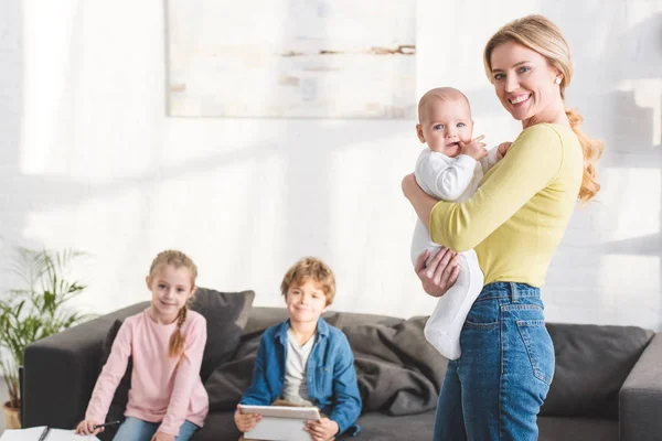 Happy mother holding infant child and smiling at camera while cute siblings sitting on sofa behind — Stock Photo