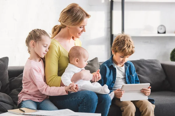 Schöne glückliche Mutter mit drei anmutigen kleinen Kindern, die zu Hause zusammen auf dem Sofa sitzen — Stockfoto