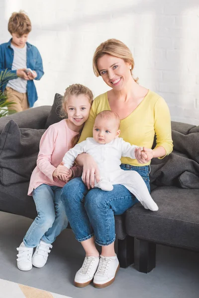 Glückliche Mutter mit Säugling und entzückender Tochter, die auf der Couch sitzt und in die Kamera lächelt — Stockfoto