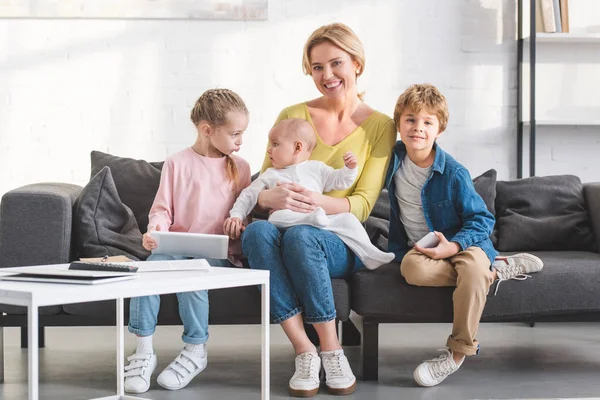 Felice madre sorridente alla macchina fotografica mentre seduto sul divano con tre adorabili bambini — Foto stock